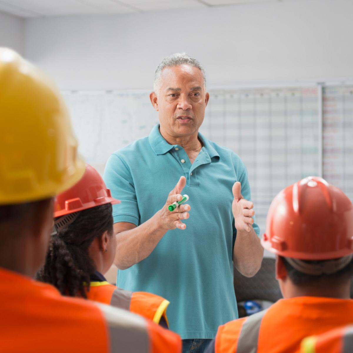 Man in blue shirt presenting to a room full of workers sitting down and wearing safety gear