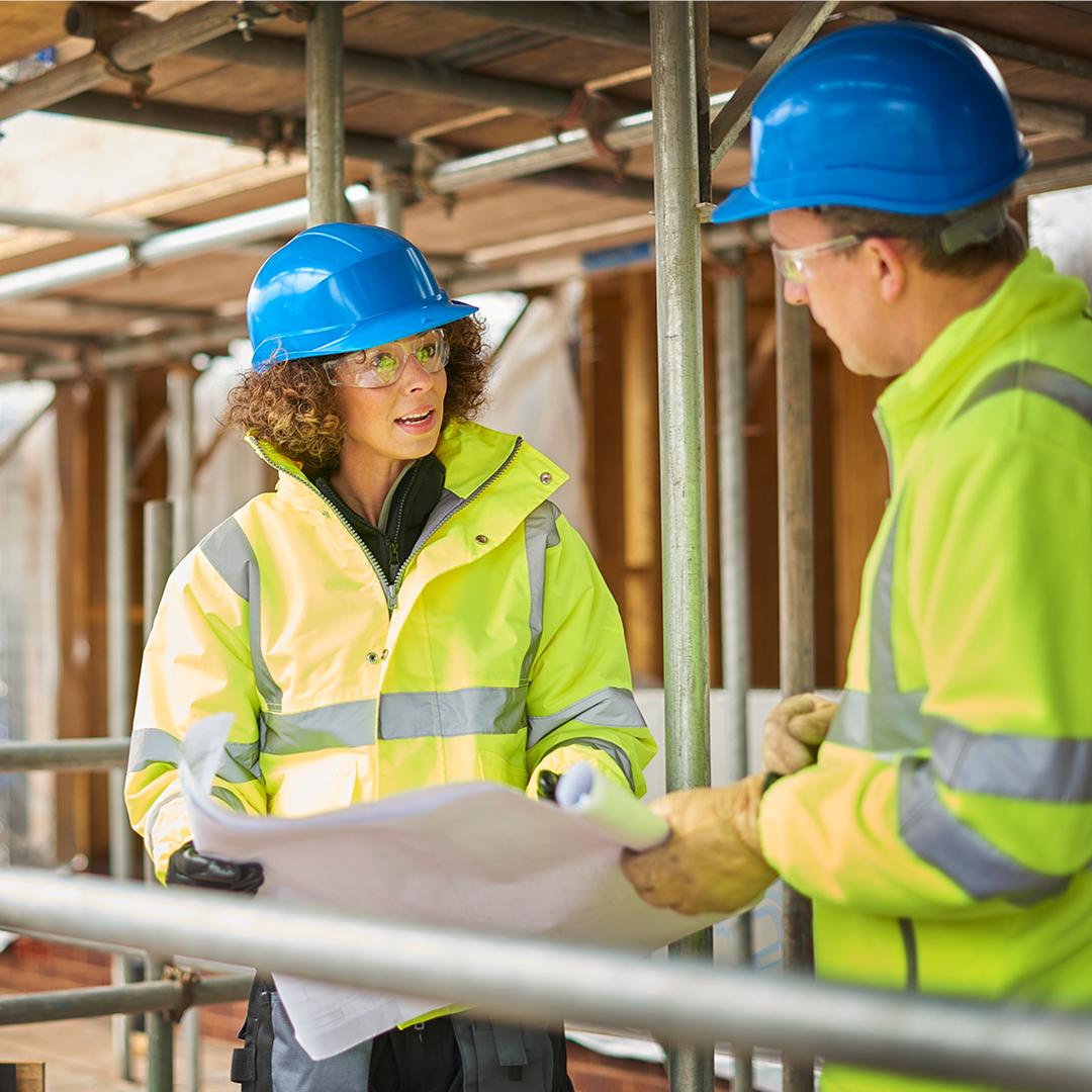 A man and woman wearing high visibility vests and hard hats engaged in conversation on a construction site